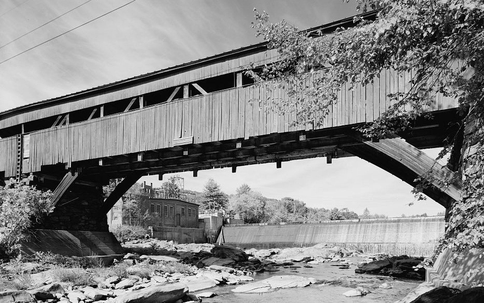 Wooden covered bridge spans a rocky river beside a low dam and small building