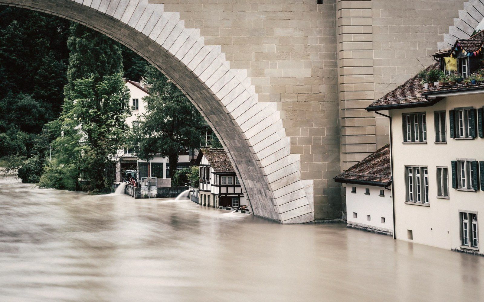 Floodwater rises beneath a stone bridge arch, submerging riverside buildings up to their windows