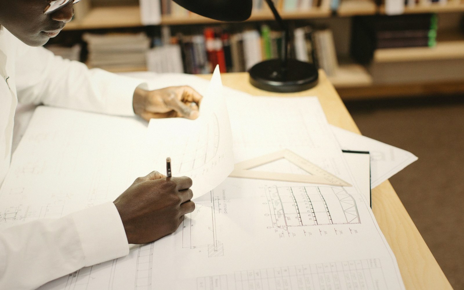 Hands holding a pencil and drawing over architectural blueprints with a triangle ruler on a desk