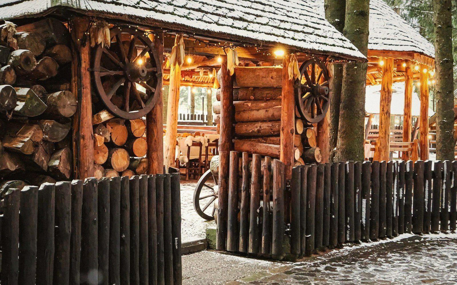 Wooden lodge entrance decorated with cart wheels, stacked firewood and string lights in snow
