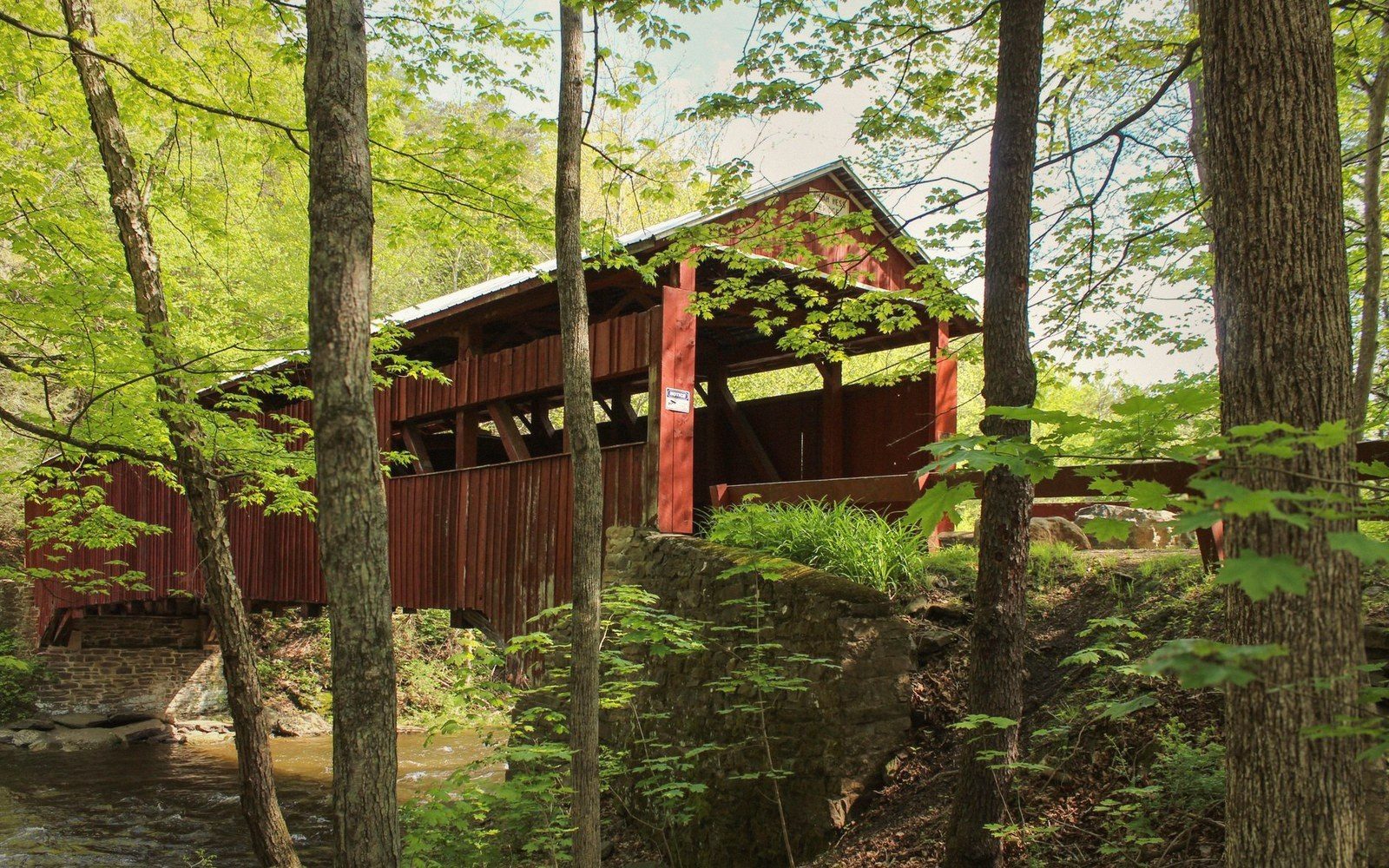 Red covered bridge spanning a creek, surrounded by dense green forest and rocky embankment