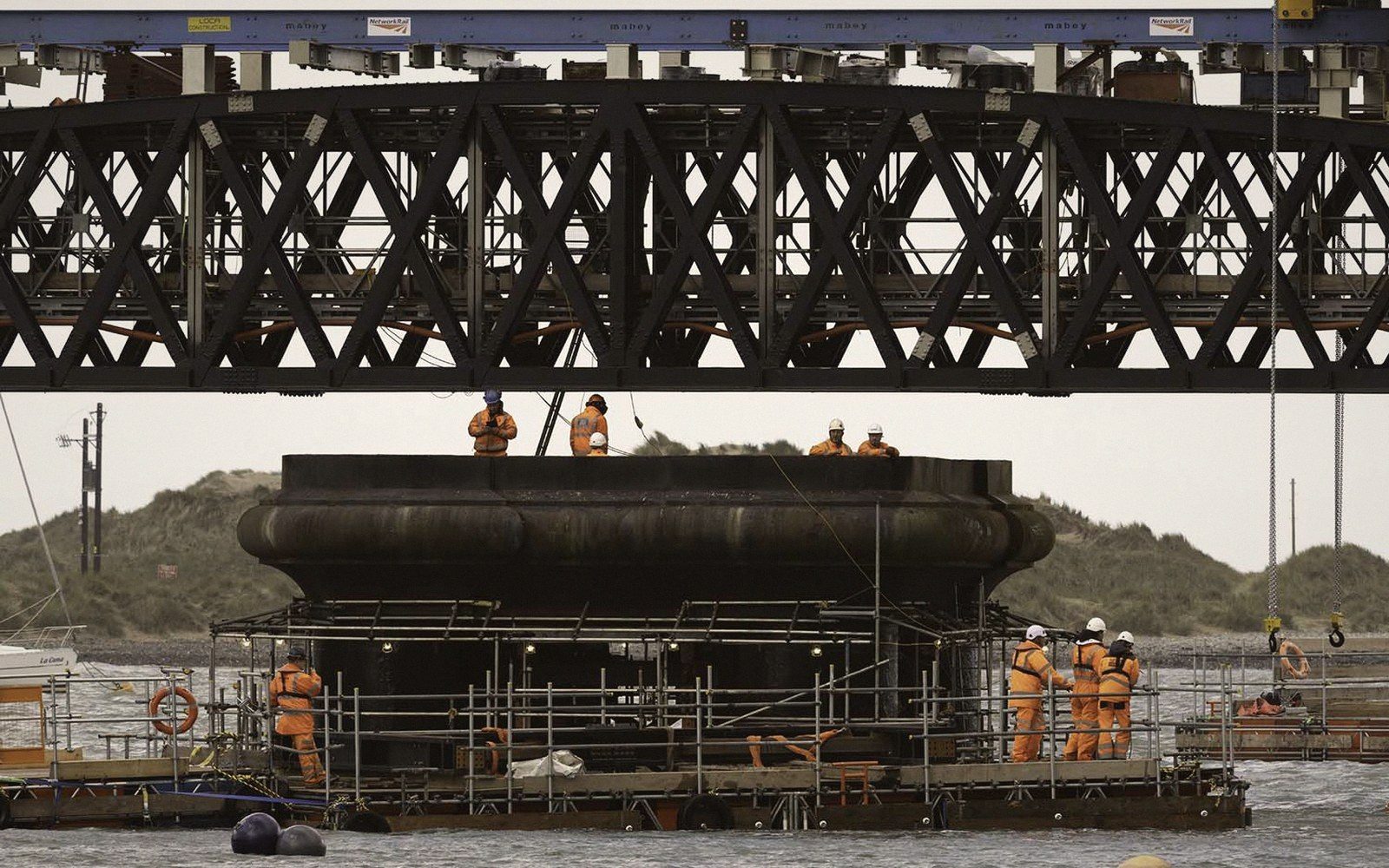 Workers in orange safety gear inspect a large bridge pier beneath a steel truss span