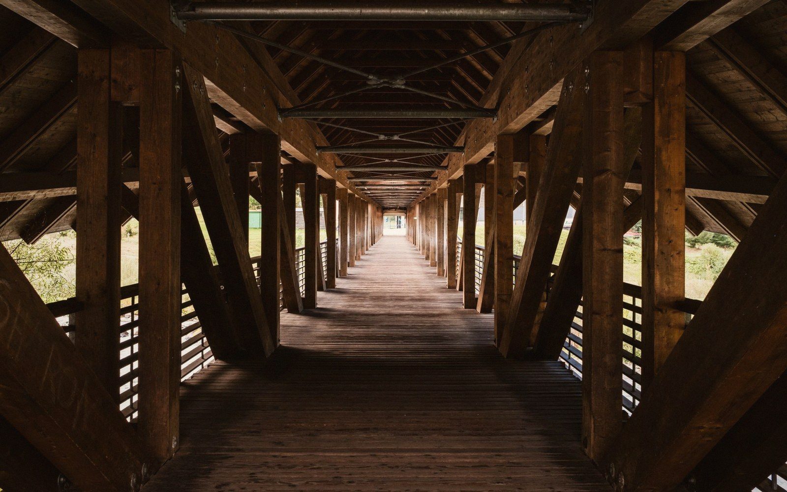 The interior of a long covered timber bridge, braced posts down both sides and the deck running away to a bright far portal