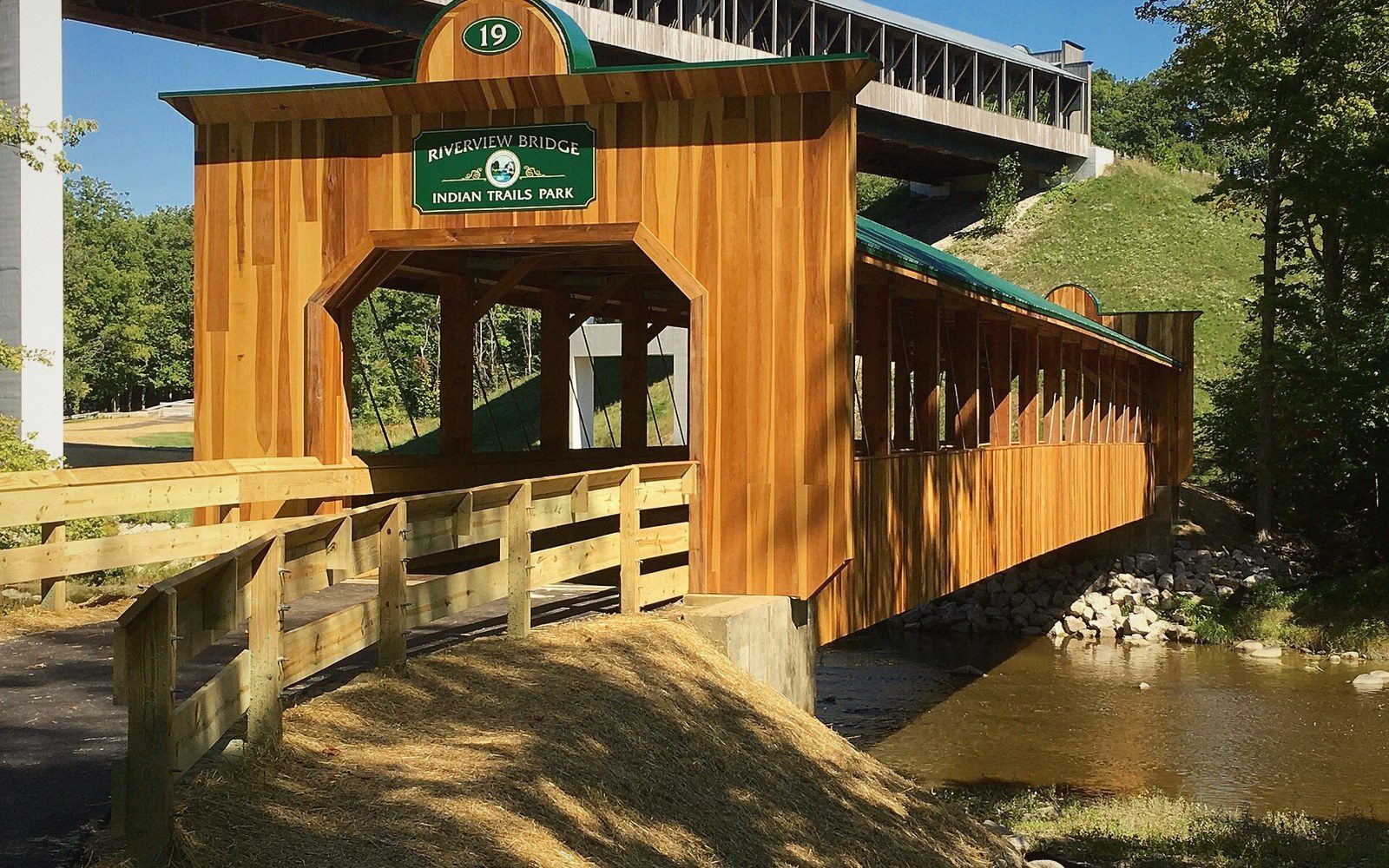 A newly built covered timber footbridge with pale cladding and a green metal roof, a concrete highway bridge behind it